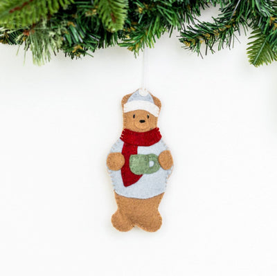 Decorative bear ornament with a scarf and mug, hanging against a white background with greenery at the top.