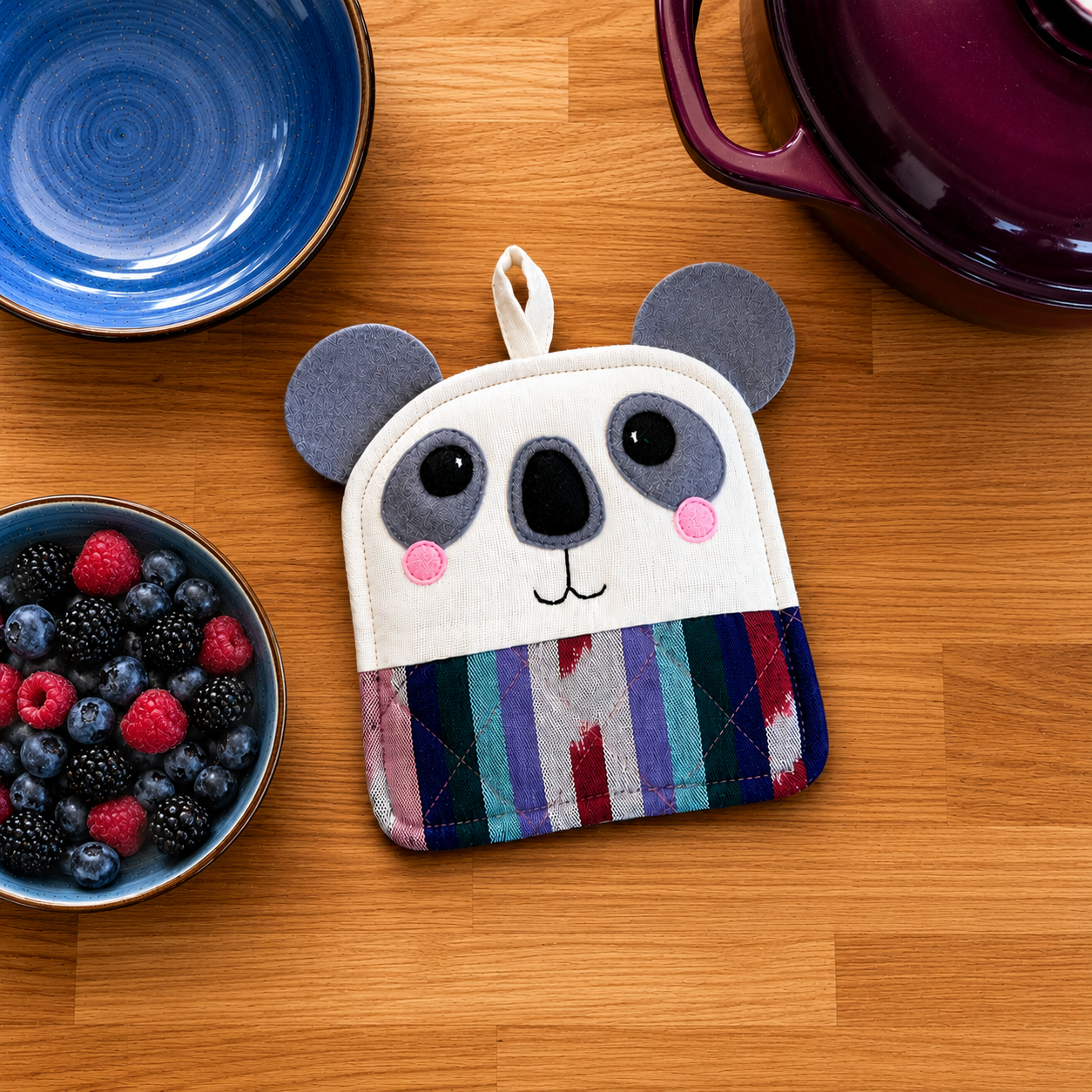 Koala shaped pot holder on a wooden surface with bowls of berries and a blue plate.